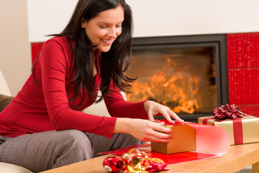 Happy woman in red wrapping Christmas present by home fireplace; 
