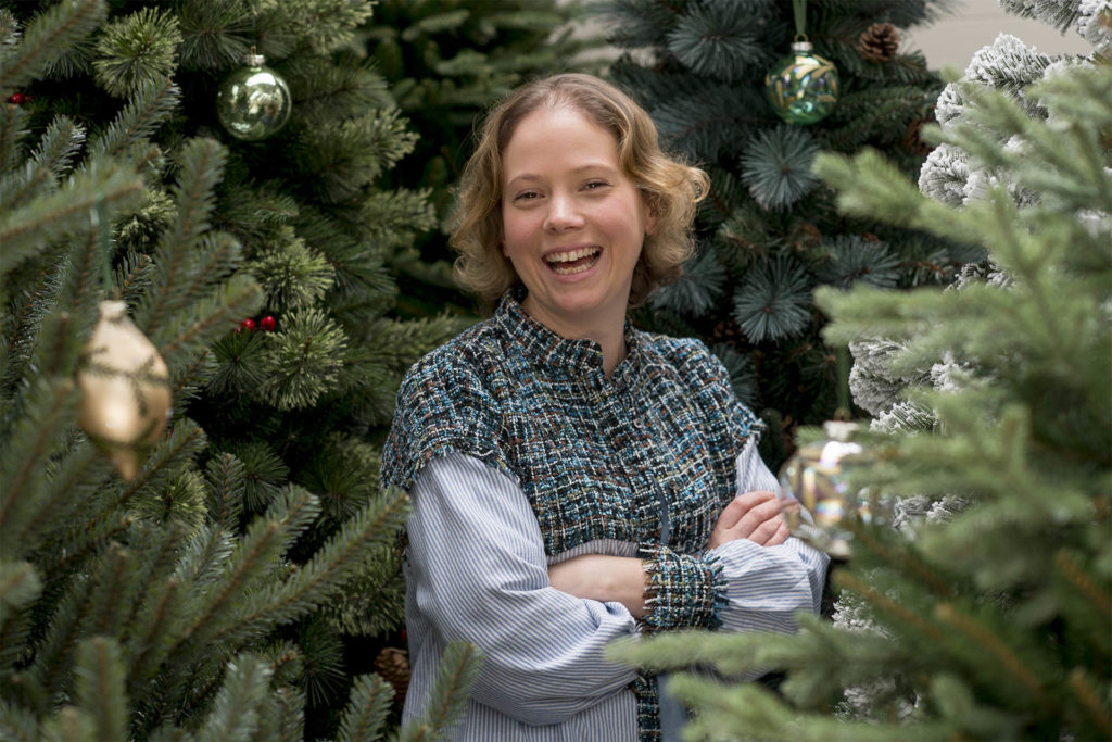 Young woman laughing, standing amongst tall real Christmas trees, few baubles