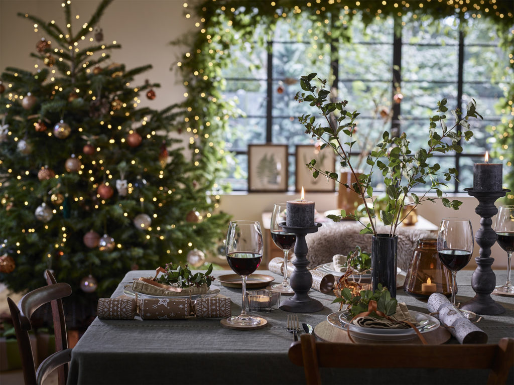 Table with candlesticks and wine glasses, large green tree and green window garland both dotted with white fairy lights